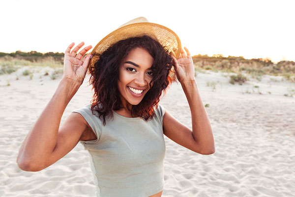 Smiling woman wearing a straw hat and a light shirt, standing on a sandy beach, representing a carefree and joyful atmosphere.