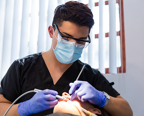 Dentist in black scrubs performing emergency dental treatment on a patient, utilizing dental tools and wearing protective gear in a well-lit office setting.