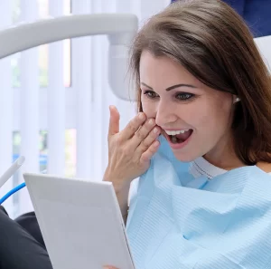 Woman in dental chair, smiling and examining her teeth on a tablet, wearing a dental bib, in a modern dental office setting, reflecting satisfaction with cosmetic dentistry results.